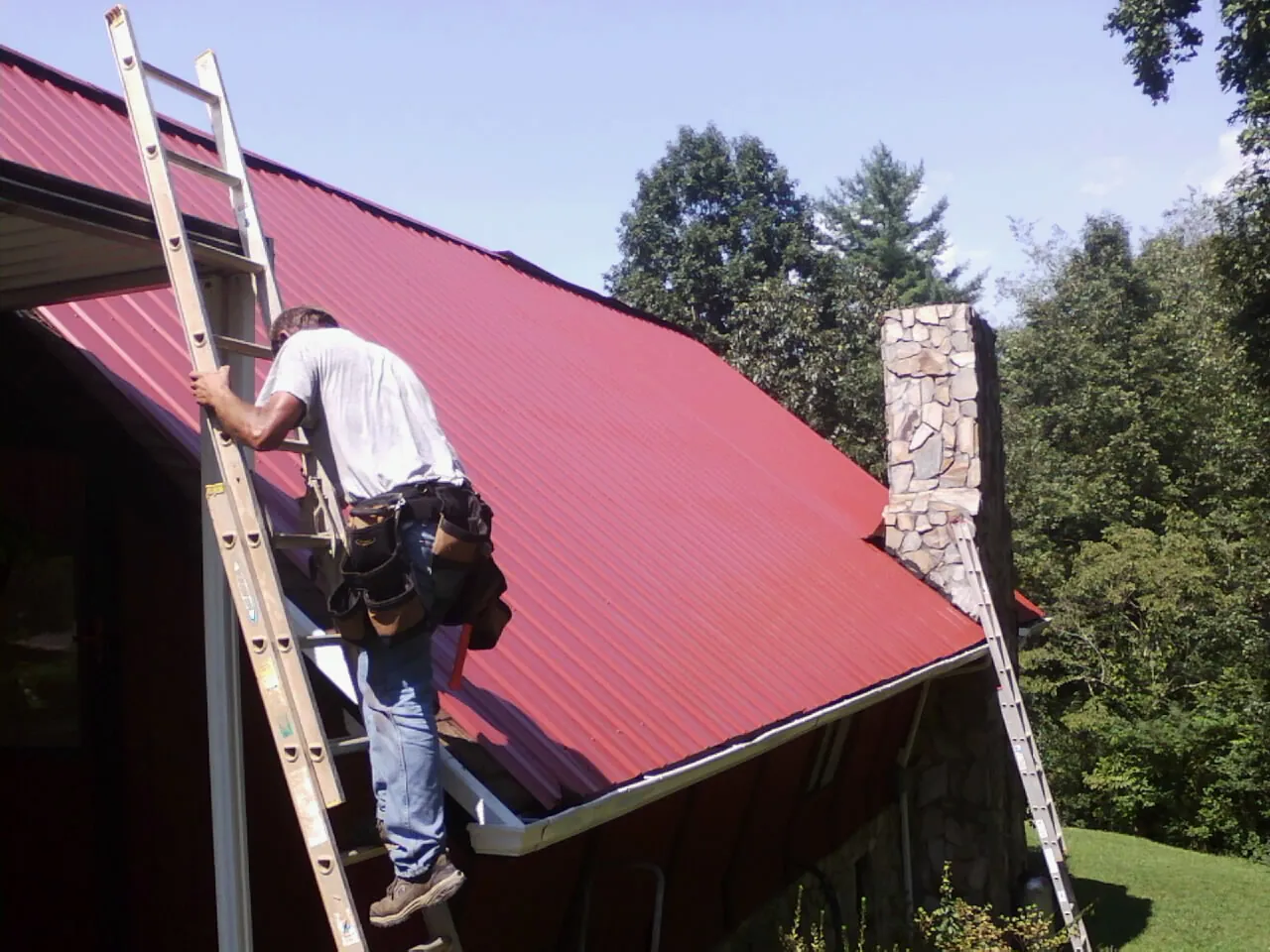 Roofer on a ladder beside a red metal roof and stone chimney — Soffit & Fascia Repair by Roofing Brothers in Louise